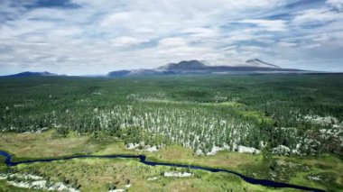 Norveç 'in dolambaçlı nehri, bereketli bitki örtüsü ve boreal çam ormanları üzerinde nefes kesici bir hava yolculuğu. Etrafı görkemli gri dağ sıralarıyla çevrili, parlak mavi gökyüzü ve bulutlar altında.