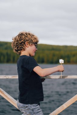 Sarışın, kıvırcık saçlı, tatlı bir çocuk göl kıyısında, akşam üstü vakti doğa tarafından çevrelenmiş, yüzünde saf bir neşe ile şiş üzerinde kızartılmış şekerlemenin tadını çıkarıyor.