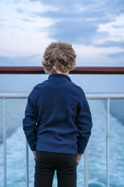 Küçük bir çocuk bir yolcu gemisinin korkuluklarında durup okyanus dalgalarını seyrediyor. Gün batımının yumuşak ışığı sakin bir atmosfer yaratır. Norveç kıyısına yakın.