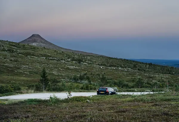 İsveç 'in Idre Dalarna şehrinde geniş bir manzara gözler önüne serilirken çakıllı bir yolda park edilmiş bir araba, alacakaranlık sırasında sakin tepeler ve uzak Stadjan dağ zirveleri sergiliyor..