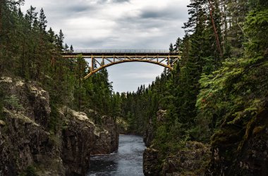 Orsa Dalarna İsveç 'te bulutlu bir gökyüzü altında sık ormanlar ve kayalık uçurumlar tarafından çerçevelenen Derin Kanyon Storstupet' te, nehirden geçen bir demiryolu köprüsü uzanır.