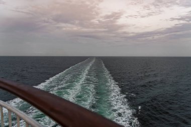 Bir yolcu gemisinin denizde dalgalanmalar bırakarak bir uyanış oluşturduğu engin okyanus manzarası. Gökyüzü alacakaranlığın yumuşak tonlarını taşıyor ve dingin atmosfere katıyor.