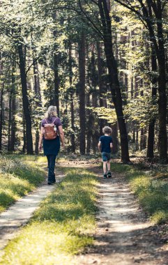 Anne ve Çocuk Huzur Ormanı 'nda Bir Yolda Yürüyorlar. Bir kadın ve küçük oğlu, uzun ağaçlarla çevrili toprak bir patikada yürüyor. Gün ışığında huzurlu bir anın tadını çıkarıyorlar.