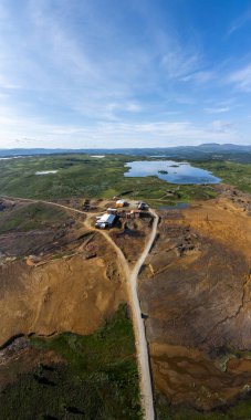 Roros Trondelag Norveç 'teki terk edilmiş tarihi bir bakır madeninin engin ve topraksı manzarasını keşfedin, çarpıcı turuncu renkler ve taze yeşilliklerle çevrili sakin mavi bir göl. Dikey Havadan Panoramik Görünüm