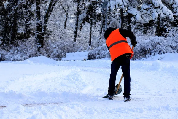 Clearing street from snow. A worker, janitor in yellow vest, cleans city street, park from fresh snow with shovel in winter. Dnipro, Ukraine
