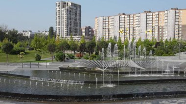 Yüksek evler, binalar ve gökdelenler karşısında fıskiyeler ve ağaçlarla dolu güzel bir park. İnsansız hava aracı manzarası, şehir manzarası. Dnipro, Ukrayna 'da bahar, yaz çeşmesi, 2021-07-01