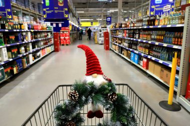 Süpermarket arabasında komik bir noel cücesi ve çelenk. Alışveriş arabası, içinde alkol, yiyecek ve yiyecek bulunan raflar. Mağazadan alışveriş, yeni yıl indirimi.