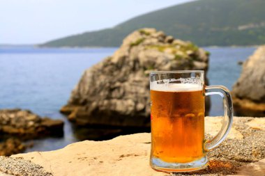 Glass of light beer near sea, close-up. Misted mug, pint of cold beer stands against background of blurred sea landscape in cafe, restaurant, bar, pub, Herceg Novi, Montenegro