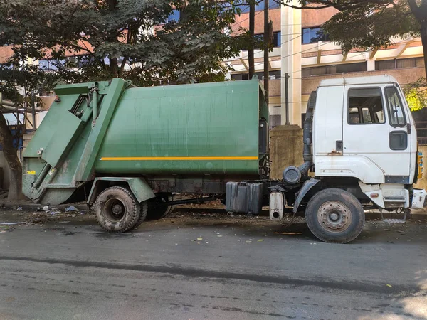Beyaz taksisi ve hidrolik kolu olan yeşil bir çöp kamyonu bir şehir caddesinde park halinde, temizlik ve sağlık hizmetlerinin korunmasında çöp kamyonlarının hayati rolünü vurguluyor..