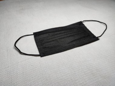 Tek kullanımlık siyah bir maske krem beyaz bir yüzey üzerinde yatar. Görüntü, yüz maskesinin basitliğini ve işlevselliğini yakalayarak, kamu sağlığı ve güvenliğindeki temel rolünü gözler önüne seriyor..