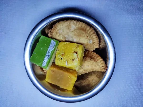 Yuvarlak metal bir kapta canlı Hint tatlıları. Sergide altın-kahverengi Soan Papdi, yeşil Besan Ladoo ve sarı Motichoor Ladoo yer alıyor..