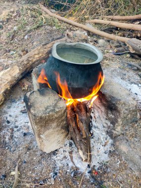 Traditional water heating with a stone-and-log charcoal stove and a large pot. The pot sits on burning charcoal, with vibrant flames heating the water, showcasing rustic simplicity and efficiency. clipart