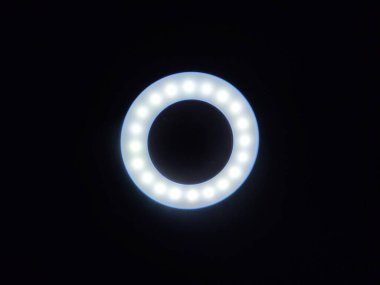 Dairesel bir LED halka ışığı, eşit aralıklı ampuller fotoğraf, video ya da içerik oluşturma için idealdir, karanlık bir arkaplan karşısında yumuşak bir şekilde parlar. Işıklandırma, teknoloji veya yaratıcılık temaları için mükemmel.