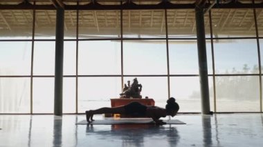 Yoga stüdyosunun geniş penceresinin önünde farklı pozisyonlarda yoga yapan tanınmayan bir kadın görülüyor. 4K Yatay Video.