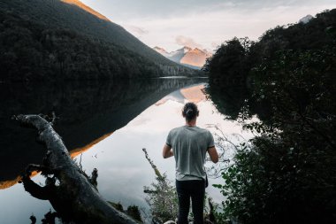 Sakin ve rahat Kafkasyalı genç adam sakin göl kenarındaki dağların ve bulutların etkileyici manzarasının güzelliğine hayran kalıyor ve hayran kalıyor ve büyük kütük, Milford ses, yeni