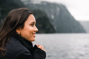 Soğuk bir günde kayalık dağların yanındaki gölün güzelliğini sakince düşünürken profili uzun saçlı beyaz kız Milford Sound, Yeni Zelanda seyahat konsepti.