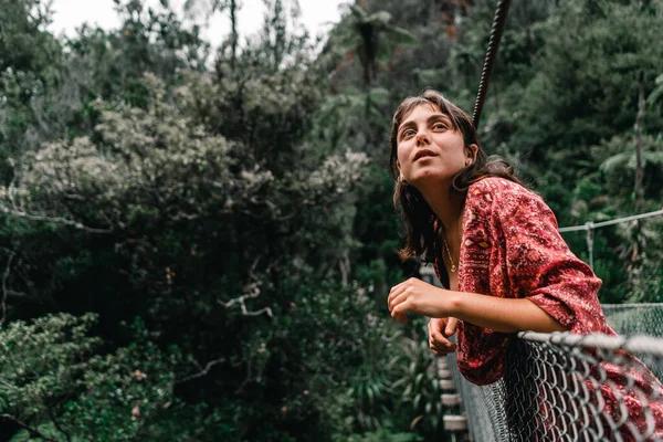 Boynunda mavi gözlü genç beyaz kadın kulaklarında yuvarlak küpeler Donut Adası, Yeni Zelanda 'daki asma köprünün korkuluklarından gökyüzüne bakıyor.