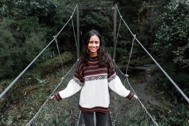 Beyaz saçlı, uzun saçlı, yün kazak giyen genç bir kadın boynunda bir gerdanlık ormanın içindeki ağaçların arasından nehri geçen metal köprüde sakin ve rahat duruyor.