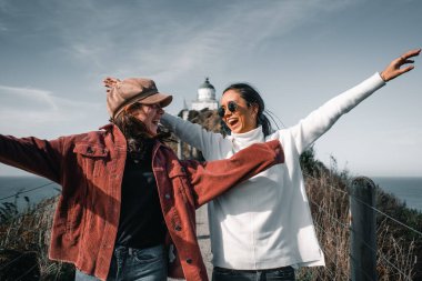 İki beyaz kız ayakta eğleniyor, mutlu ve gülümsüyor. Toprak yolda kollarını açmış, güzel beyaz deniz fenerinden geliyor. Nugget Point, Yeni Zelanda - Seyahat kavramı