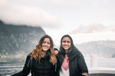 İki uzun saçlı beyaz kadın birlikte fiyort gölü ve dağı ziyaret ederek turistik güvertede eğleniyorlar, Milford Sound, Yeni Zelanda - seyahat konsepti