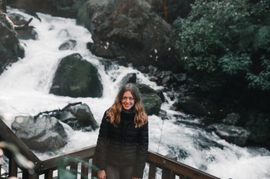 Sarışın, beyaz bir kadın kameraya gülümseyen siyah ceketli gri pantolonlu, bitkilerin ve ormanların ortasındaki etkileyici şelalenin yanındaki ahşap pervaza yaslanmış.