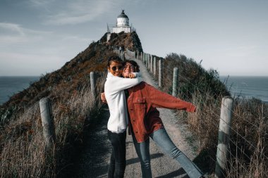 couple of brunette caucasian girls disheveled by the wind embracing happily having fun on the path that leads to the impressive lighthouse, nugget point, new zealand - Travel concept clipart