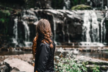 Kahverengi saçlı, saçları örgülü, siyah ceketli güzel şelaleleri seyreden sakin ve rahat şelalelere, purakaunui şelalelerine, Yeni Zelanda 'ya seyahat eden beyaz kız.