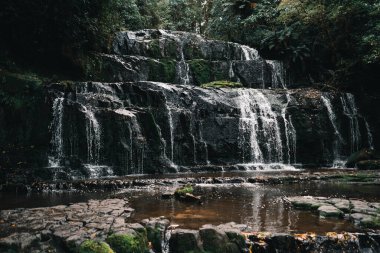 Güzel şelaleler büyük kayaların arasından dökülüyor. Küçük gölün yakınındaki orman ağaçlarının arasından. Doğanın sessizliğinde bir sürü taş var. -