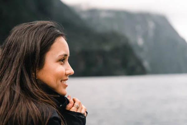 Uzun kahverengi gözleri ve siyah ceketi olan, dağların ve ormanın yanındaki gölün güzelliğini, Milford Sound 'u, Yeni Zelanda' yı düşünüp rahatlatan beyaz tenli genç bir kadın.