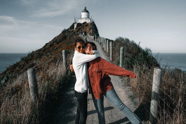 Rüzgarla darmadağın olmuş birkaç esmer beyaz kız etkileyici deniz fenerine, Nugget Point 'e ve Yeni Zelanda' ya giden yolda mutlu mesut kucaklaşıyorlar.