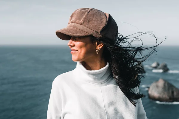 stock image brunette caucasian girl in profile with nice white sweater hat black hair moved by the force of the wind near the sea and rocks smiling calm, nugget point, new zealand - Travel concept