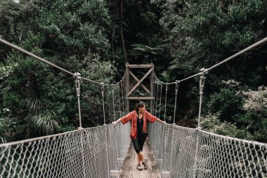 Mavi gözlü, dağınık saçlı, siyah elbiseli, kırmızı ceketli, uzun ve dar bir köprünün korkuluklarına tutunmuş beyaz bir kız. Yeni Zelanda 'nın Çörek Adası' ndaki nehre bakıyor. -