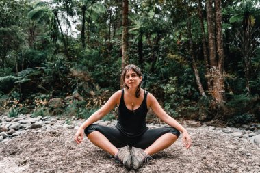 Dağınık mavi gözlü, yuvarlak küpeli ve boynunda kolyesi olan beyaz bir kız kameraya bakıyor. Kollarını dizlerine uzatmış bir şekilde Çörek Adası 'ndaki ormanda oturuyor.