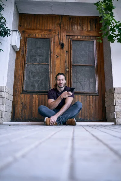 caucasian young man with long hair and beard with smartphone in hand sitting by wooden door of house looking at camera - Urban style