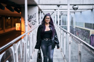 Uzun saçlı, mavi pantolonlu, elinde çantasıyla tren istasyonuna giden yolda yürüyen esmer, genç bir kadın.