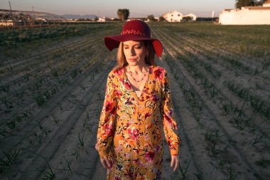 Gün batımında ekilmiş bir tarlanın ortasında dikilirken, çiçekli bir elbise giymiş, sarışın, beyaz, olgun bir kadın.