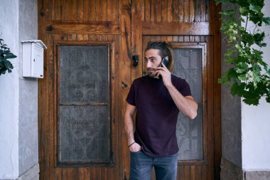 Beyaz, uzun saçlı ve sakallı bir genç, akıllı telefonuyla konuşuyor. Evin ahşap kapısının yanında gülümsüyor. Elleri cebinde.