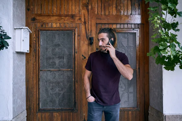Caucasian young man with long hair and beard talking on the smartphone of him smiling near the wooden door of the house with his hand in his pocket - Urban style
