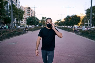 Uzun saçlı, beyaz bir adam bulvarda yürüyor akıllı telefondan gülümsüyor ve şehirde yüzünde maskeyle kameraya bakıyor.