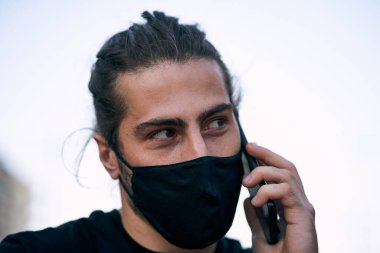 Kafkasyalı adam siyah tişört dağınık uzun saçlı, maskeli, şehirde akıllı telefonuyla konuşan biri.