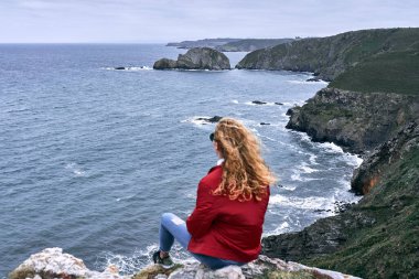 Sarışın, beyaz, kırmızı ceketli ve mavi pantolonlu bir kız sakin bir kayanın üzerinde oturmuş deniz dalgalarının güzelliğine hayran, Cabo de Penas, İspanya manzarası