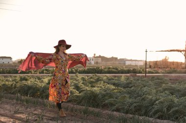 Yetişkin, beyaz bir kadın, sakin bir şekilde bahçenin içinde dolaşıyor, pembe bir şalla büyük kırmızı bir şapka takıyor ve kollarını açıyor.