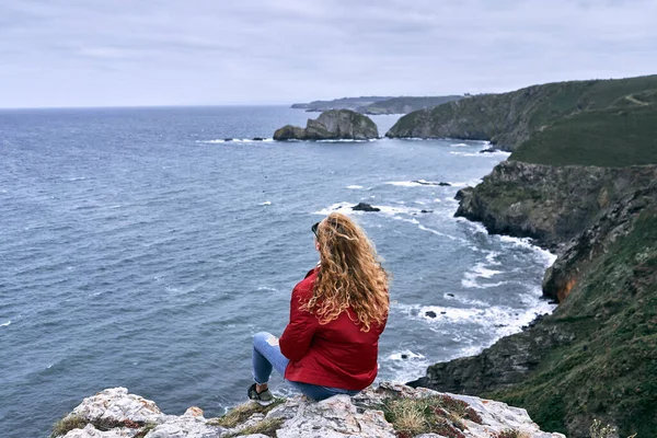 Sarışın, beyaz, kırmızı ceketli ve mavi pantolonlu genç bir kadın kayalıklarda sakin ve deniz dalgaları, Cabo de Penas, İspanya manzaraları ile rahatlamış bir şekilde oturuyor.