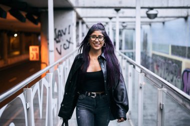 Uzun saçlı esmer, mavi pantolonlu genç Latin kadın tren istasyonuna giden yolda yürürken kameraya gülümsüyor.