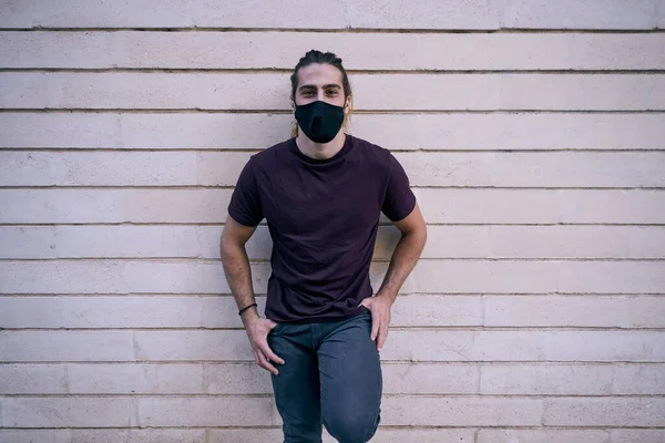 caucasian young man in black t-shirt and face mask with thumbs in pockets and crouched left leg standing by wall - Urban style