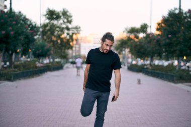 Beyaz adam siyah tişört darmadağın olmuş uzun saçlı, sakallı sağ ayağını tutuyor ve elini şehir bulvarında caddede duruyor.