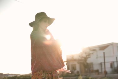 Pembe şallı ve büyük kırmızı şapkalı beyaz bir kadın güneşli bir günde arka plana bakıyor.
