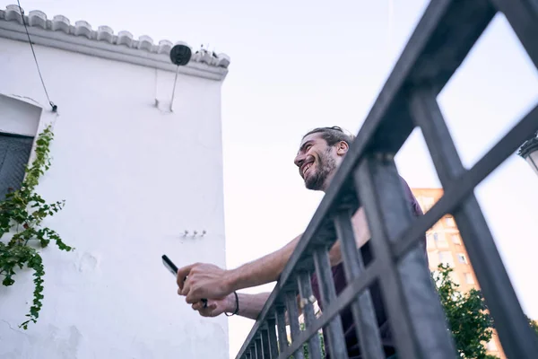 Kafkasyalı, uzun saçlı, mutlu, pis bir korkuluğa yaslanmış, akıllı telefonunun ekranına bakan genç bir adam.