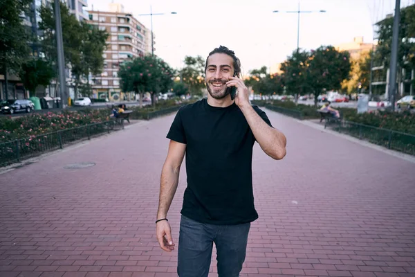 stock image young caucasian man black t-shirt beard looking at camera smiling talking on the smartphone standing on the boulevard in the city - Urban style