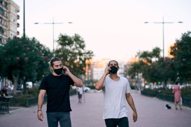 İki beyaz tişörtlü ve kot pantolonlu genç adam sokakta yan yana yürüyorlar ve yüz maskeli akıllı telefonlarıyla konuşuyorlar.